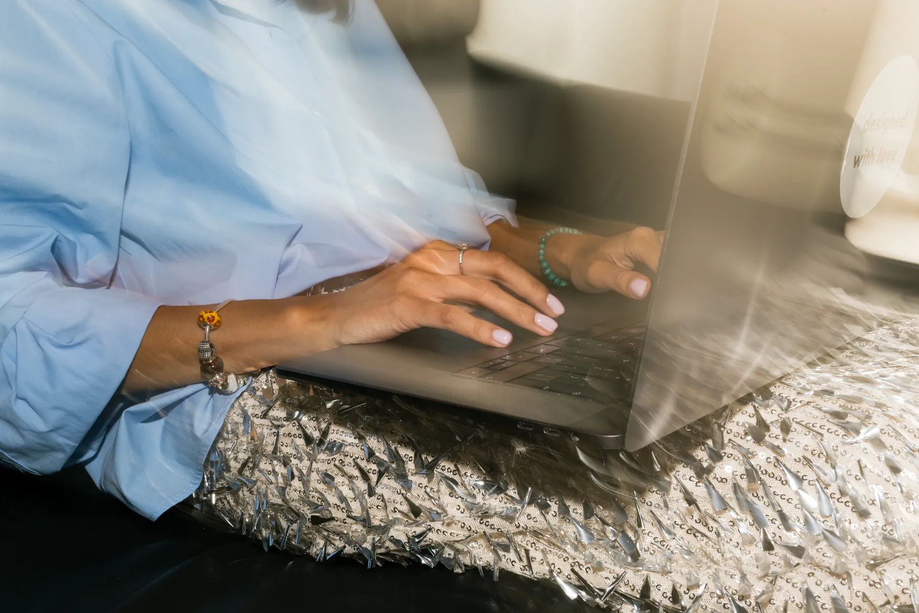 Hands typing on a laptop, seen through a window, the desk below catching the light off a sequinned surface