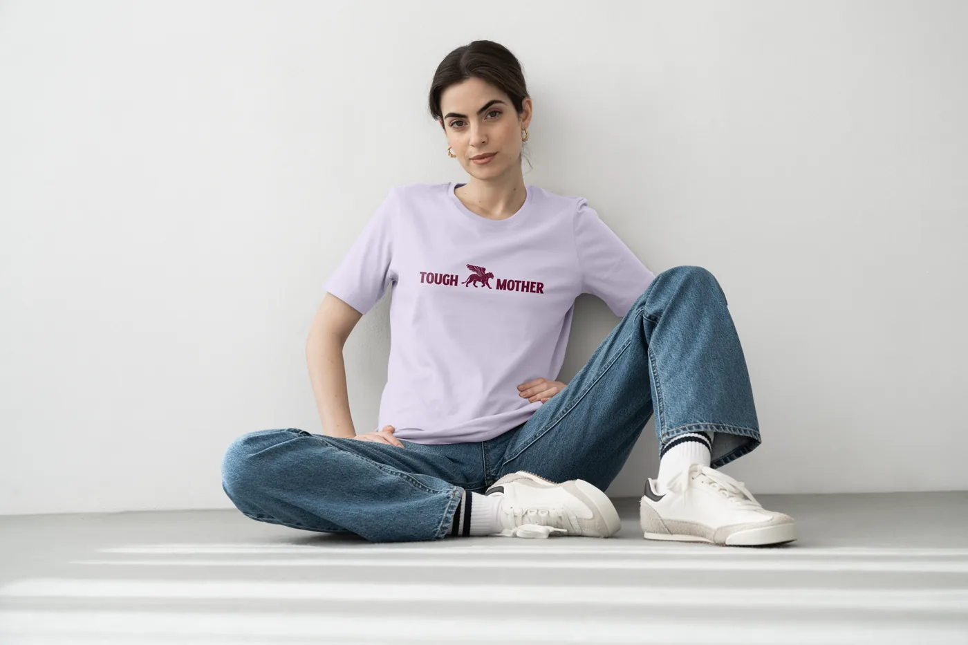 A woman in a lilac Tough Mother t-shirt, sitting against a white wall