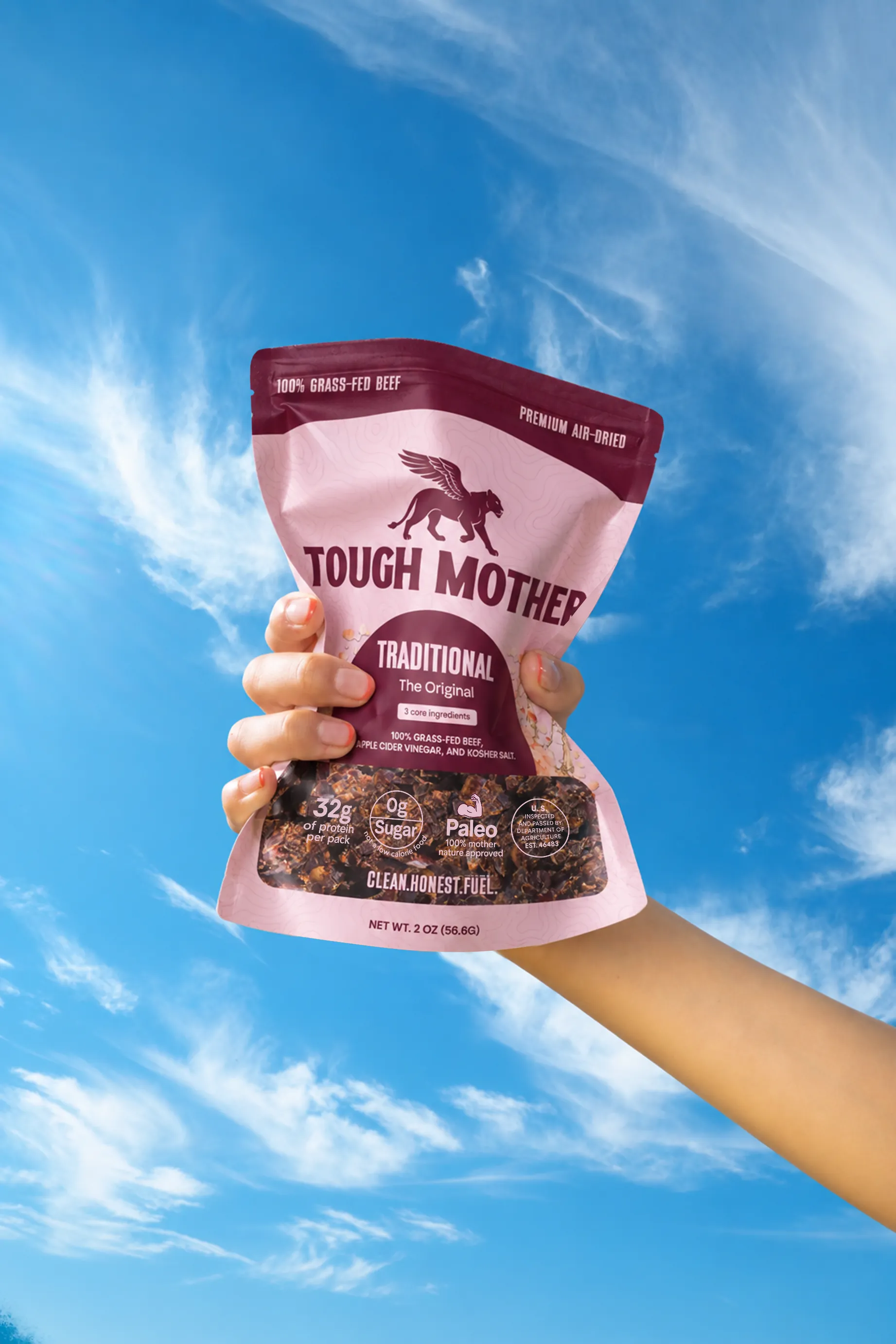 A hand holding up a pouch of Tough Mother biltong against a blue sky