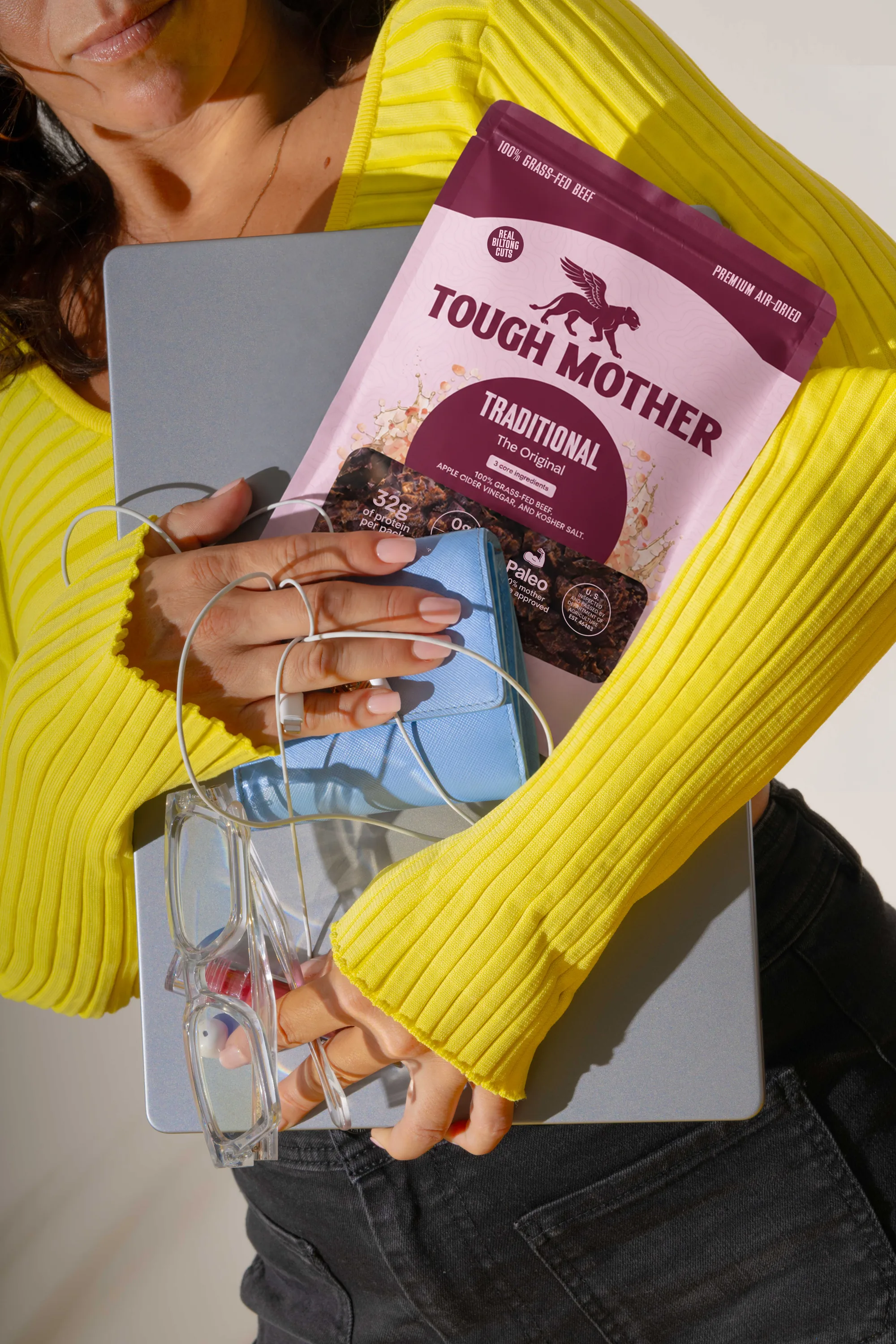 A woman in a yellow sweater holding a Traditional pouch against a laptop and a pair of glasses